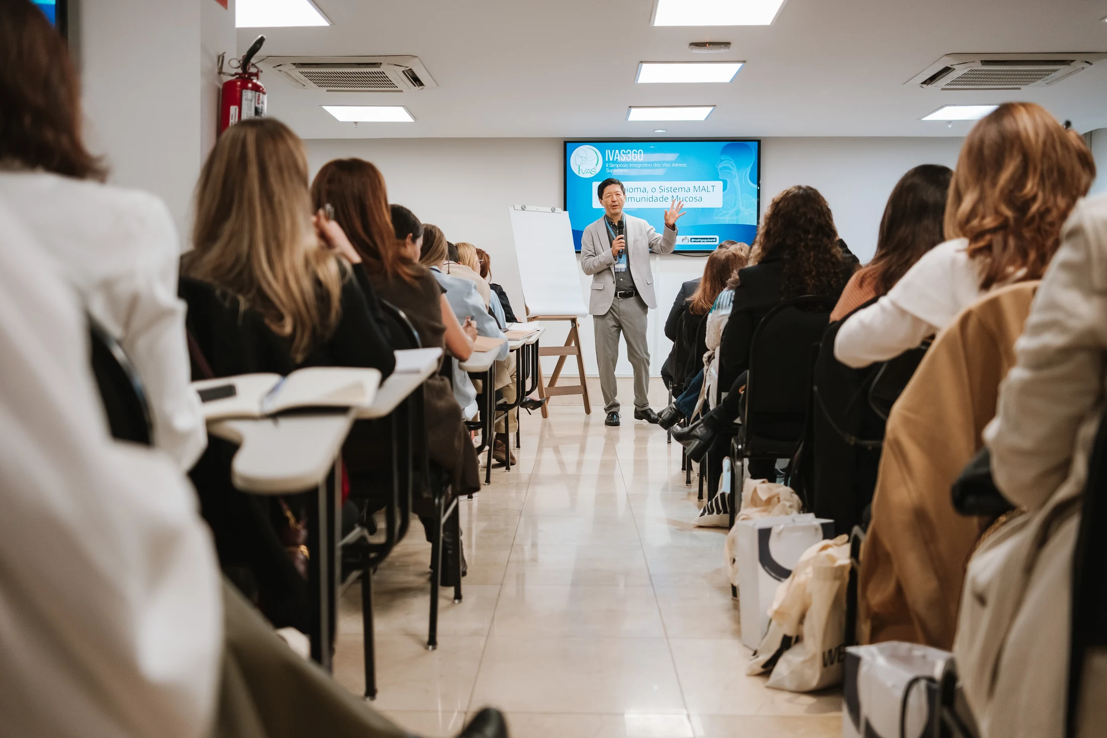 Dr. Milton Takeuti conduzindo uma aula presencial para médicos, com os participantes sentados dos dois lados do corredor